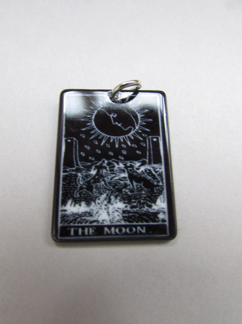May include: Black and white tarot card pendant with a silver bail. The card depicts the Moon card from the tarot deck. The card features a crescent moon, two towers, a dog, a crab, and a path. The text "THE MOON" is printed at the bottom of the card.