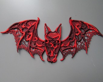 Puede incluir: Un parche bordado con forma de murciélago que presenta un intrincado trabajo de hilo rojo y negro. El diseño muestra un patrón detallado similar al encaje en las alas y una cabeza estilizada, creando una silueta simétrica con reflejos rojos.