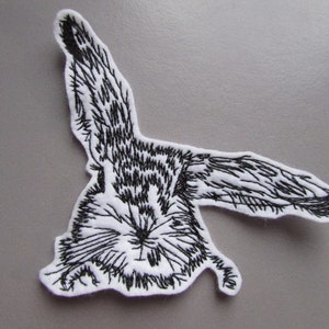 May include: Black and white embroidered patch of a rabbit with its ears up. The rabbit is lying on its side with its head turned to the side.