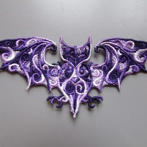 May include: Purple and white embroidered bat applique with intricate details. The bat has a detailed body and wings with a swirling pattern.