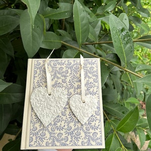 Puede incluir: Dos adornos de cerámica blanca en forma de corazón con un diseño floral cuelgan de un diario de color crema con un patrón floral azul.