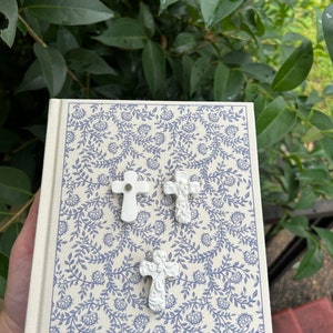 May include: Three white ceramic cross charms with intricate designs on a cream-colored notebook with a blue floral pattern.