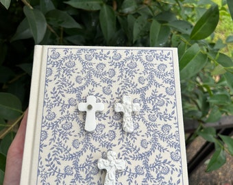May include: Three white ceramic cross charms with intricate designs on a cream-colored notebook with a blue floral pattern.