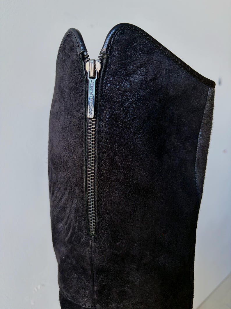May include: Close-up of a black suede boot with a silver zipper. The zipper runs vertically along the side of the boot. The brand name "DONALD PLINER" is visible on the zipper pull. The boot has a sleek, modern design.