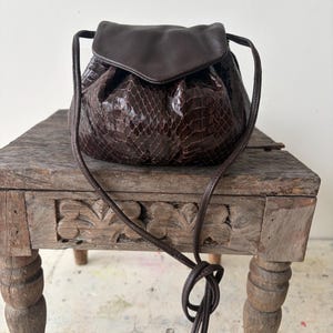 May include: A brown leather handbag with a snakeskin pattern. The bag has a flap closure and a long, thin strap. The bag is sitting on a wooden table.