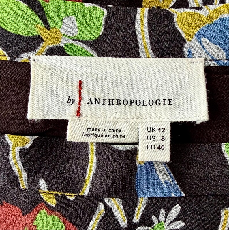 Peut inclure: Gros plan sur une &eacute;tiquette de v&ecirc;tement avec la marque "ANTHROPOLOGIE" en noir. L'&eacute;tiquette indique les tailles : UK 12, US 8, et EU 40. Le tissu en dessous pr&eacute;sente un motif floral avec des touches de vert, jaune, bleu et rouge.