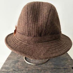 May include: A brown wool hat with a structured crown and a wide brim. The hat features a decorative feather and is displayed on a silver stand. The hat has a textured surface and a classic design.