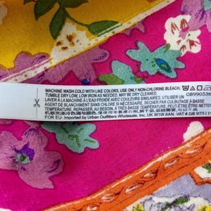 May include: A fabric label with care instructions, set against a colourful floral background. The label is white with black text, and the background features pink, yellow, and orange floral patterns. Washing instructions and a product code are visible.