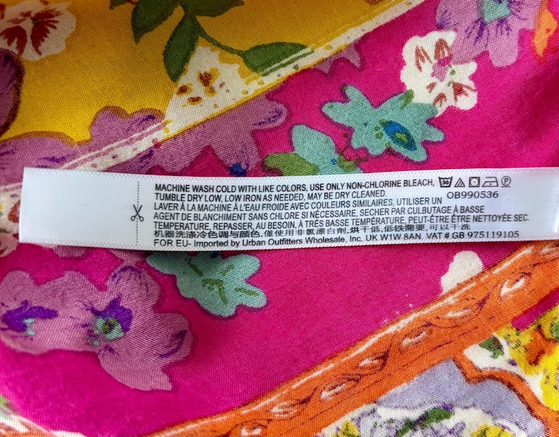 May include: A fabric label with care instructions, set against a colourful floral background. The label is white with black text, and the background features pink, yellow, and orange floral patterns. Washing instructions and a product code are visible.