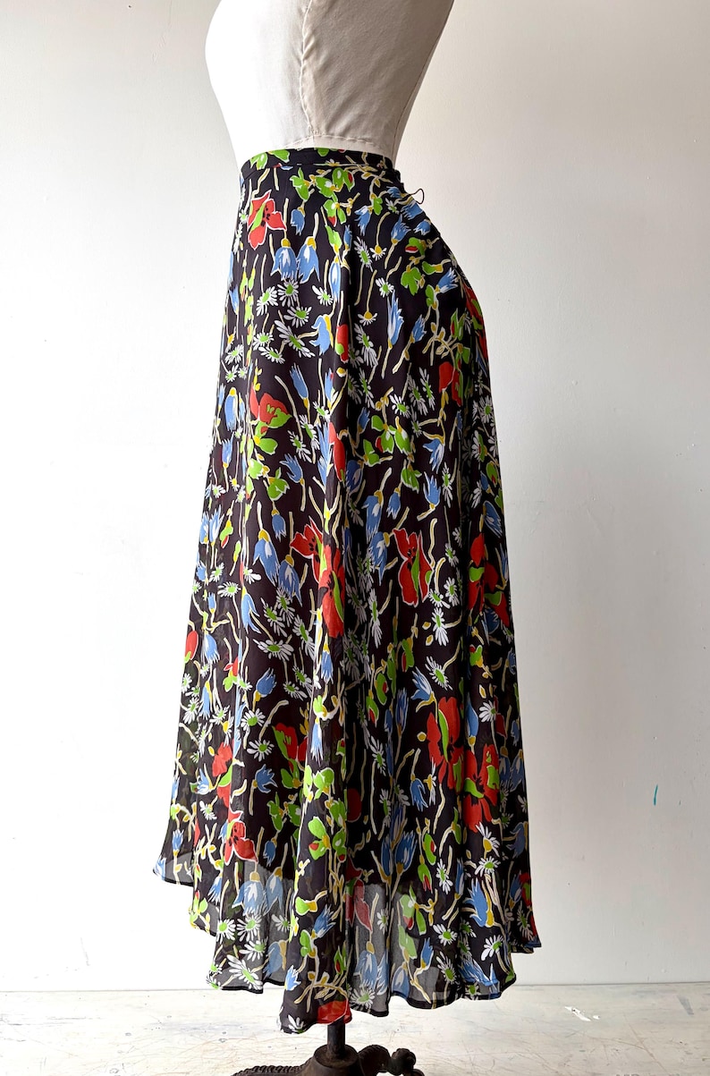 Peut inclure: Longue jupe fluide avec un fond sombre et un motif floral vibrant. Le motif pr&eacute;sente des fleurs rouges, bleues et vertes avec des accents blancs. La jupe est pr&eacute;sent&eacute;e sur un mannequin.
