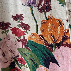 K&ouml;nnte beinhalten: Ein strukturierter, cremewei&szlig;er Stoff mit einem lebendigen Blumenmuster. Das Design zeigt gro&szlig;e orangefarbene, rosafarbene und dunkelrote Bl&uuml;ten mit dunkelgr&uuml;nen Bl&auml;ttern und St&auml;ngeln. Die Webstruktur des Materials ist mit horizontalen Linien sichtbar.