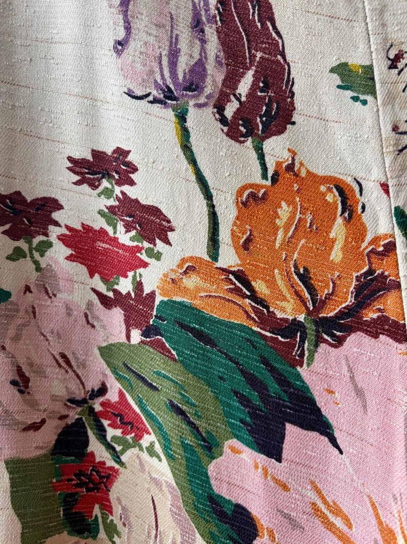K&ouml;nnte beinhalten: Ein strukturierter, cremewei&szlig;er Stoff mit einem lebendigen Blumenmuster. Das Design zeigt gro&szlig;e orangefarbene, rosafarbene und dunkelrote Bl&uuml;ten mit dunkelgr&uuml;nen Bl&auml;ttern und St&auml;ngeln. Die Webstruktur des Materials ist mit horizontalen Linien sichtbar.