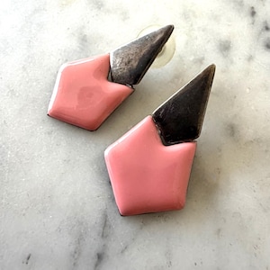 May include: A pair of geometric earrings. Each earring features a light pink, pentagon-shaped base with a dark, triangular top. The earrings have a post backing for pierced ears. The earrings are made of metal.