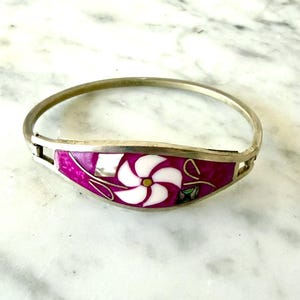 May include: A silver-toned hinged bangle bracelet featuring a central curved panel inlaid with vibrant magenta enamel, a white five-petaled flower design, iridescent mother-of-pearl accents, and thin silver-colored metal lines throughout.