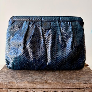May include: A dark blue clutch bag with a snakeskin pattern. The rectangular bag has a gathered design and a small, dark leather accent. The bag is on a wooden table.