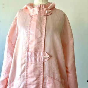 May include: A light pink hooded jacket featuring a front zipper closure concealed by a placket with two horizontal tabs secured by small, textured, light-colored toggles. The jacket includes long sleeves and two front pockets with flap closures.