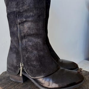 May include: Black knee-high boots with a side zipper. The boots have a leather appearance with a suede-like upper section. The boots are displayed on a wooden surface. The boots have a low heel.