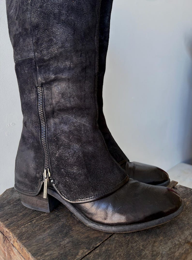 May include: Black knee-high boots with a side zipper. The boots have a leather appearance with a suede-like upper section. The boots are displayed on a wooden surface. The boots have a low heel.