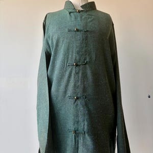 May include: A dark green long-sleeved tunic with a mandarin collar and frog closures. The tunic is made of a textured fabric and is displayed on a mannequin. The tunic has a relaxed fit and a classic design.