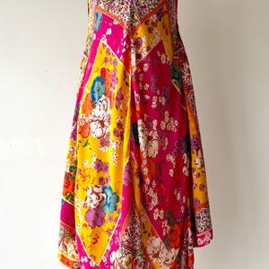 May include: A colourful patterned dress with a V-neck and thin straps. The dress has a patchwork design with bright pink, yellow, and floral patterns. The dress is displayed on a mannequin against a white background.