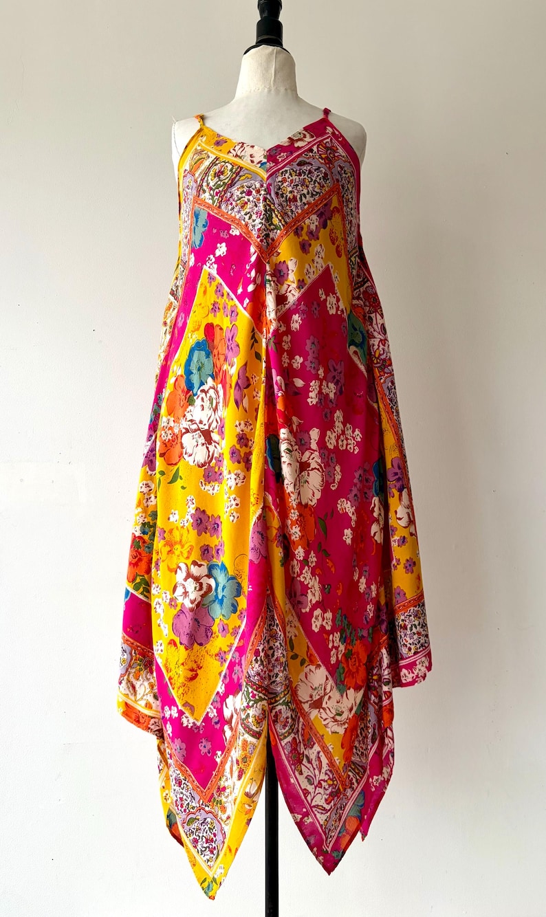 May include: A colourful patterned dress with a V-neck and thin straps. The dress has a patchwork design with bright pink, yellow, and floral patterns. The dress is displayed on a mannequin against a white background.
