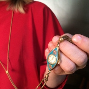 May include: A gold chain necklace with a vintage, eye-shaped pocket watch pendant. The watch face is white with black hands and Roman numeral markings. The watch case is blue with pink floral accents.