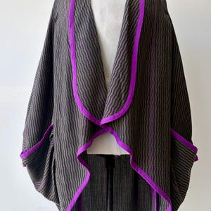 May include: A dark gray and black striped cape with vibrant purple trim. The cape features a flowing design with a wide collar and pockets. The edges and seams are accented with a bright purple fabric.