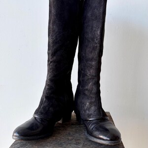 May include: A pair of black knee-high boots with a textured suede upper and a leather lower section. The boots have a zipper closure and are displayed on a wooden surface.