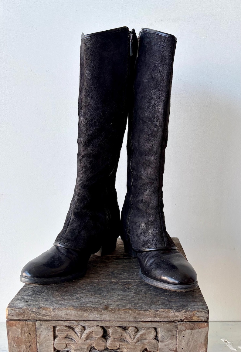 May include: A pair of black knee-high boots with a textured suede upper and a leather lower section. The boots have a zipper closure and are displayed on a wooden surface.