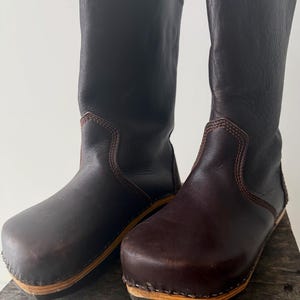 May include: A pair of dark brown leather boots with a wooden sole. The boots have a tall shaft and a rounded toe. The stitching is visible in a lighter brown color. The boots are displayed on a wooden surface.