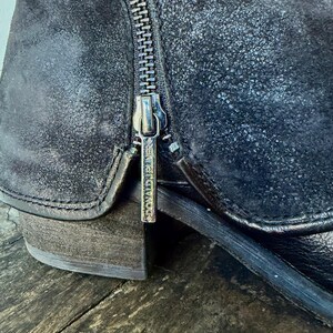 May include: Close-up of a black leather boot with a side zipper. The boot has a distressed, textured appearance and a small heel. The zipper pull has the brand name "DONALD J PLINER" engraved on it. The boot is on a wooden surface.