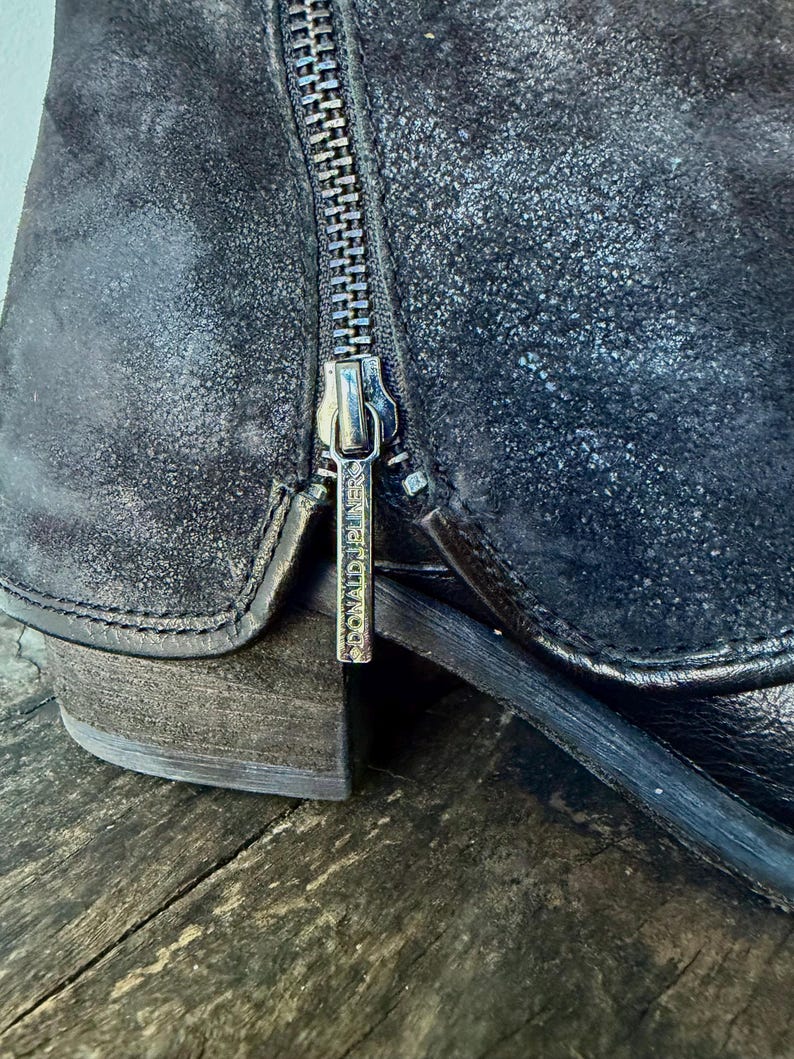 May include: Close-up of a black leather boot with a side zipper. The boot has a distressed, textured appearance and a small heel. The zipper pull has the brand name "DONALD J PLINER" engraved on it. The boot is on a wooden surface.