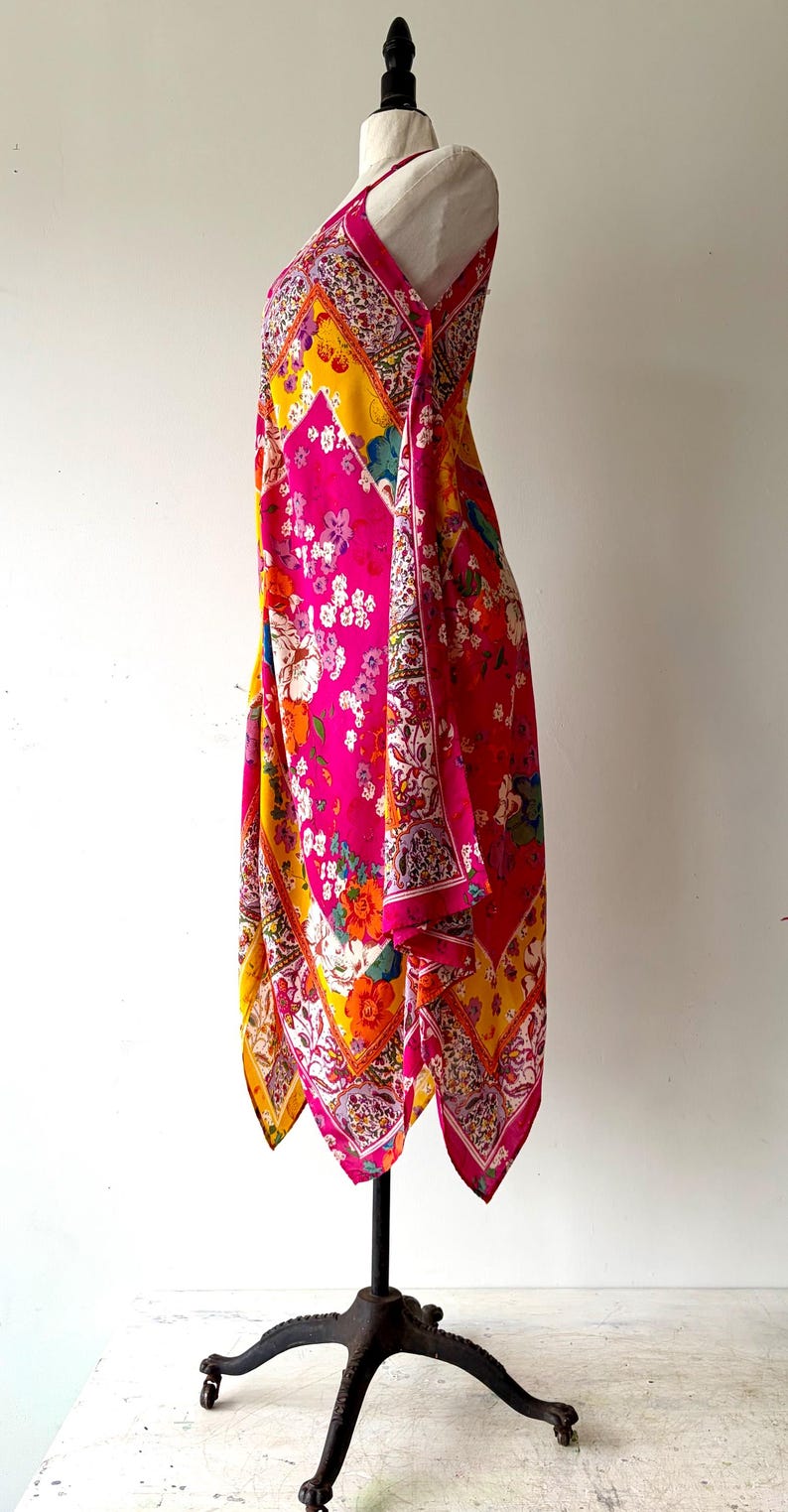 May include: A colourful patterned dress in pink, yellow, and red, displayed on a black mannequin. The dress features a floral design with a mix of colours and patterns. The dress is sleeveless and has a flowing, asymmetrical hemline.