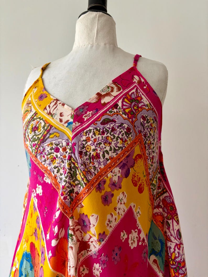 May include: A patterned dress with a V-neck and spaghetti straps. The dress features a patchwork design with vibrant pink, yellow, and floral patterns. The dress is displayed on a mannequin.