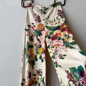 K&ouml;nnte beinhalten: Hoch taillierte Hose mit weitem Bein aus cremefarbenem Stoff mit einem lebendigen, mehrfarbigen Blumenmuster in Rot-, Rosa-, Gelb- und Gr&uuml;nt&ouml;nen. Die Hose hat einen strukturierten Bund und eine strukturierte Oberfl&auml;che und h&auml;ngt an zwei Kleiderb&uuml;geln.