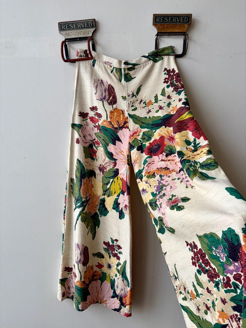 K&ouml;nnte beinhalten: Hoch taillierte Hose mit weitem Bein aus cremefarbenem Stoff mit einem lebendigen, mehrfarbigen Blumenmuster in Rot-, Rosa-, Gelb- und Gr&uuml;nt&ouml;nen. Die Hose hat einen strukturierten Bund und eine strukturierte Oberfl&auml;che und h&auml;ngt an zwei Kleiderb&uuml;geln.