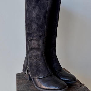 May include: A pair of black knee-high boots with a side zipper. The boots have a combination of leather and suede textures. They are displayed on a rustic wooden surface, showcasing their design and materials.