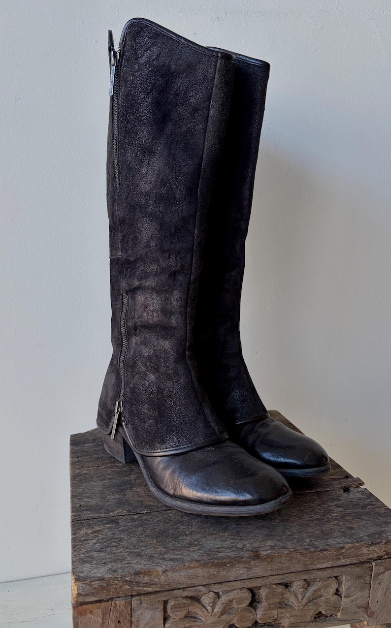 May include: A pair of black knee-high boots with a side zipper. The boots have a combination of leather and suede textures. They are displayed on a rustic wooden surface, showcasing their design and materials.