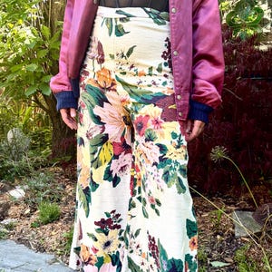 K&ouml;nnte beinhalten: Eine mauvefarbene Satin-Bomberjacke mit Druckknopfleiste, ein schwarzes Oberteil im Korsett-Stil und eine hoch taillierte Hose mit weitem Bein auf cremefarbenem Grund mit einem lebhaften Blumenmuster in Rosa, Gelb, Gr&uuml;n und Dunkelrot, kombiniert mit schwarzen Plateauschuhen.
