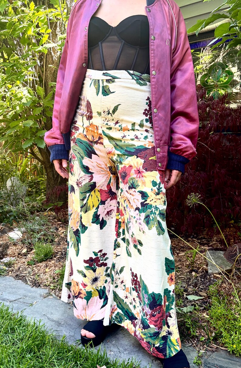 K&ouml;nnte beinhalten: Eine mauvefarbene Satin-Bomberjacke mit Druckknopfleiste, ein schwarzes Oberteil im Korsett-Stil und eine hoch taillierte Hose mit weitem Bein auf cremefarbenem Grund mit einem lebhaften Blumenmuster in Rosa, Gelb, Gr&uuml;n und Dunkelrot, kombiniert mit schwarzen Plateauschuhen.