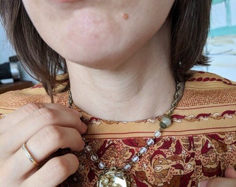 May include: A vintage-style necklace with a round, ornate pendant featuring a floral design. The pendant is made of metal and has a white background with pink and gold accents. The necklace is made of a chain with clear beads and a brown bead.
