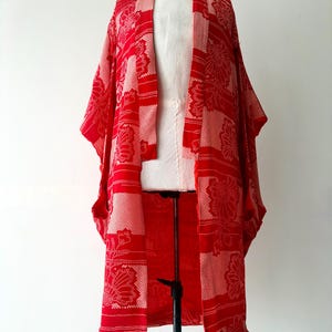 May include: A red and white patterned kimono-style robe displayed on a mannequin. The robe features a floral design and a loose, open front. The fabric has a textured appearance, and the color scheme is predominantly red and white.