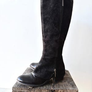 May include: A pair of black knee-high boots with a combination of suede and leather. The boots have a side zipper and are displayed on a rustic wooden surface. The boots have a pointed toe and a small heel.