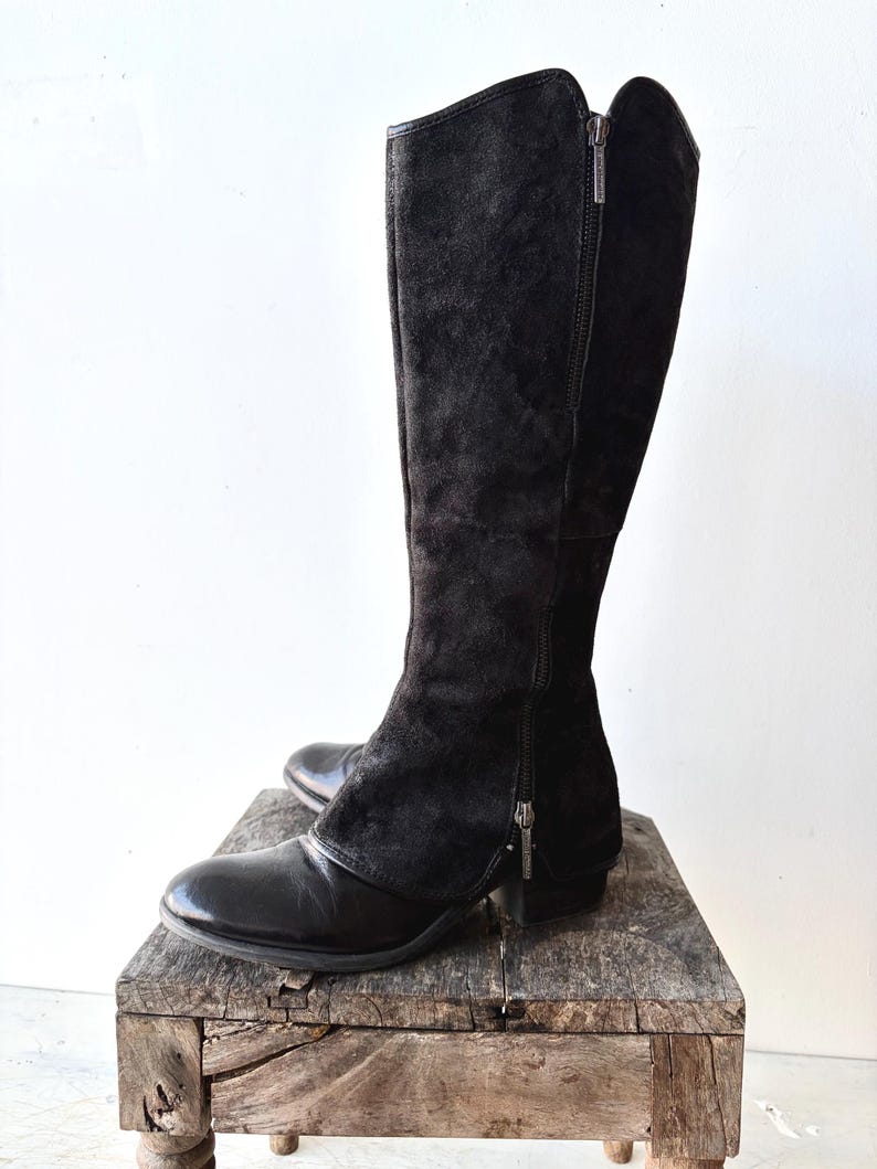 May include: A pair of black knee-high boots with a combination of suede and leather. The boots have a side zipper and are displayed on a rustic wooden surface. The boots have a pointed toe and a small heel.