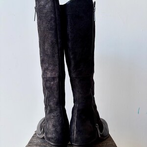 May include: A pair of black knee-high boots with side zippers. The boots are made of a textured material, possibly suede or leather, and have a low heel. The boots are standing upright on a wooden surface against a white background.