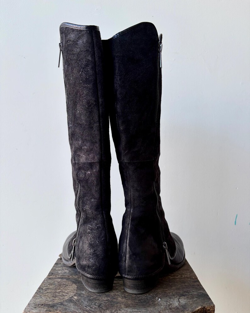 May include: A pair of black knee-high boots with side zippers. The boots are made of a textured material, possibly suede or leather, and have a low heel. The boots are standing upright on a wooden surface against a white background.