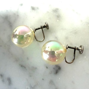 May include: A pair of vintage screw-back earrings. Each earring has a large, iridescent, round bead with a pearlescent sheen. The metal components are silver-toned, set against a white marble surface.