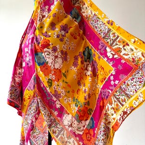 May include: A colourful, square scarf with a floral design. The scarf displays a mix of yellow, pink, orange, and white. The pattern includes large flowers and smaller floral details, with a decorative border. The scarf is made of a lightweight material.