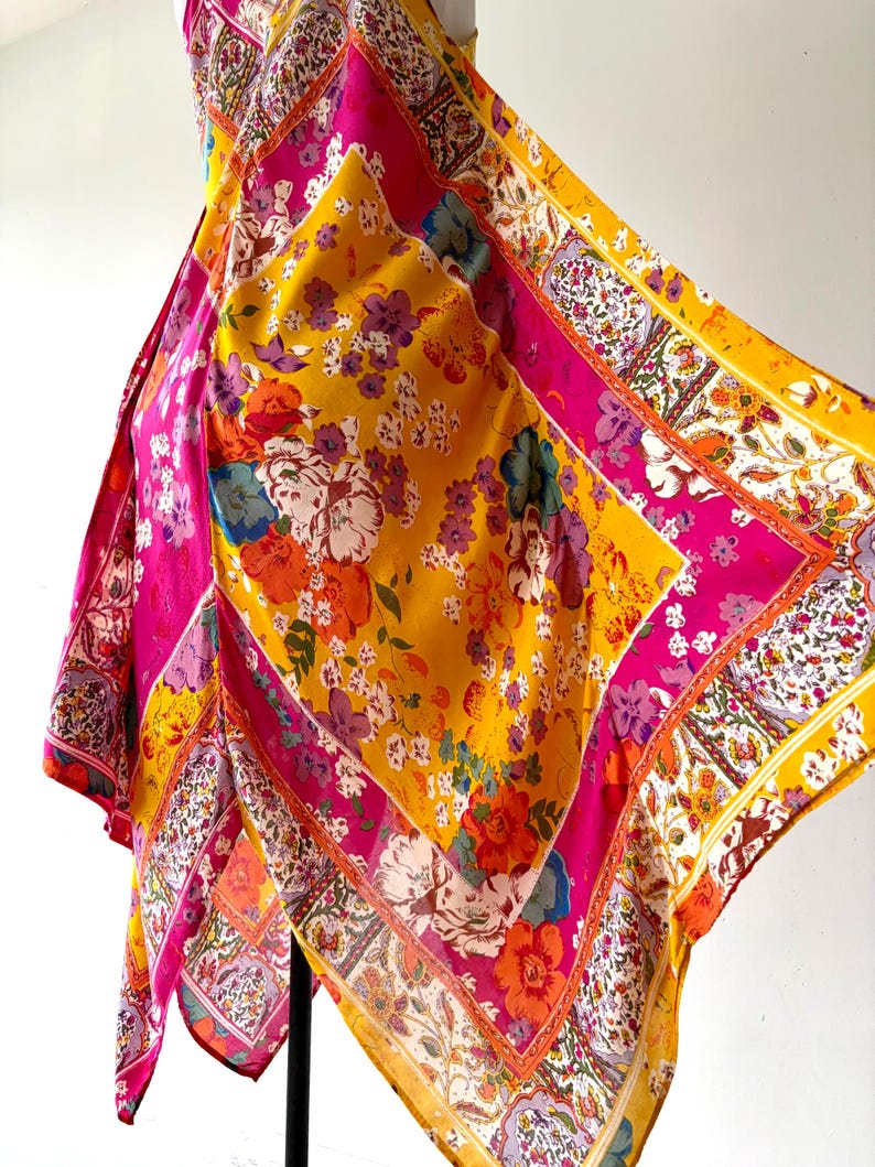 May include: A colourful, square scarf with a floral design. The scarf displays a mix of yellow, pink, orange, and white. The pattern includes large flowers and smaller floral details, with a decorative border. The scarf is made of a lightweight material.