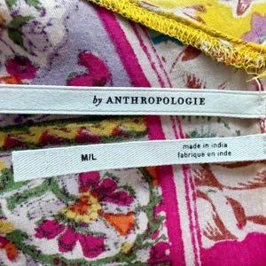May include: A close-up of patterned fabric in pink, yellow, and purple with floral designs. Two white labels are visible, one with "by ANTHROPOLOGIE" and the other with "M/L" and "made in india / fabrique en inde".
